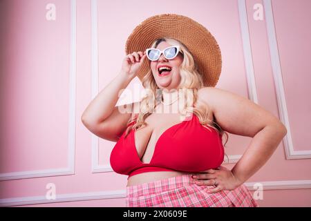 Modèle féminin excité avec sourire dentelé portant chapeau et lunettes de soleil debout derrière fond rose dans le studio Banque D'Images
