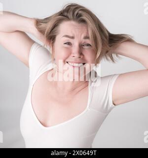 Portrait d'une femme adulte mature avec les mains derrière la tête et le sourire dentelé. La femme heureuse porte un body. La prise de vue en grand angle capture parfaitement la blonde Wom Banque D'Images