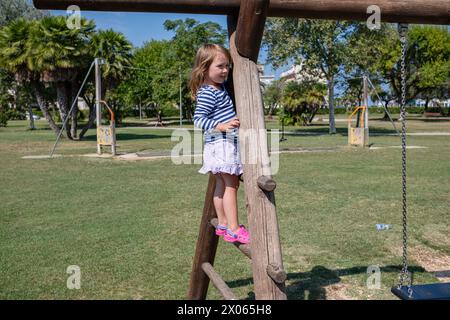Une petite fille mignonne se promène sur le terrain de jeu par une journée d'été ensoleillée. La jeune fille monte sur un toboggan, un élastique, et grimpe sur un toboggan. Banque D'Images
