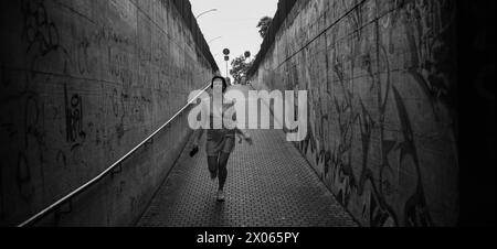 Les murs en béton du passage souterrain avec des graffitis entrent en perspective, le ciel du coucher du soleil et une fille qui court dans une robe. artis noir et blanc Banque D'Images