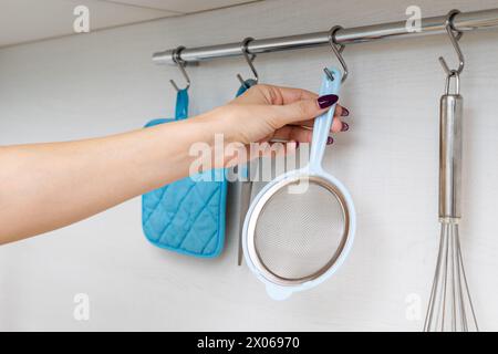 la femme au foyer retire la crépine de cuisine du crochet sur le rail. rail de cuisine avec ustensiles. tamis métallique de cuisine. Banque D'Images