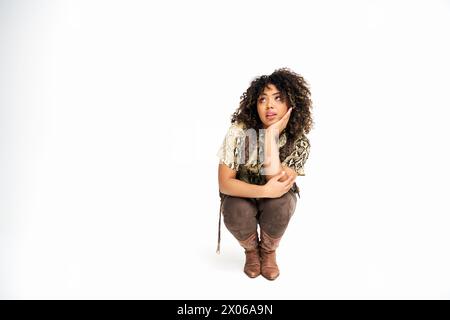 femme afro-américaine séduisante dans une tenue élégante squattant sur fond blanc et regardant loin Banque D'Images