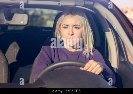 Concentrée jeune femme blonde portant un pull violet conduisant une voiture, avec une expression concentrée et légèrement tendue, suggérant une navigation prudente o Banque D'Images