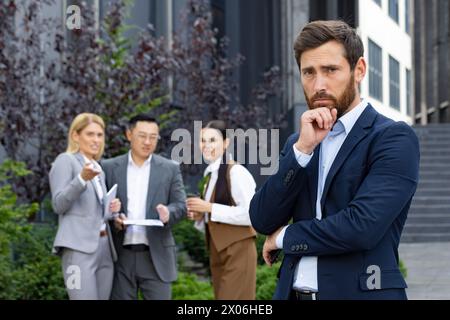 Portrait d'un jeune homme bouleversé en costume d'affaires debout à l'extérieur de l'immeuble de bureaux regardant déçu de la caméra, derrière un groupe de personnes interraciales acclamant et riant du nouveau collègue. Banque D'Images