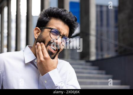 Photo en gros plan d'un jeune Indien en chemise blanche debout près d'un bâtiment, tenant son visage avec sa main et souffrant d'un mal de dents. Banque D'Images