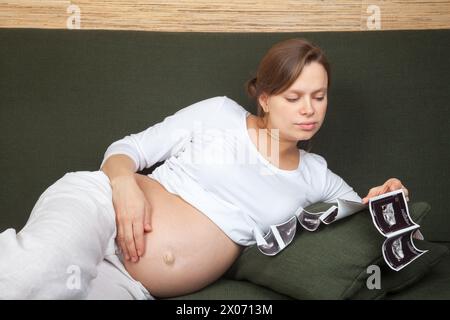 Une femme enceinte vêtue de vêtements confortables allongée sur un canapé vert examinant les échographies en train de réfléchir à son futur enfant Banque D'Images