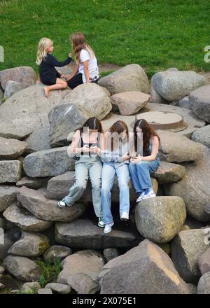 Jeunes adolescentes adolescentes avec des smartphones portant des jeans socialiser et parler assis sur des rochers dans un parc public, Gdansk, Pologne Banque D'Images