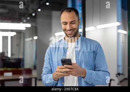 Homme hispanique indien en tenue décontractée, heureux en regardant son téléphone dans un cadre de bureau contemporain. Banque D'Images