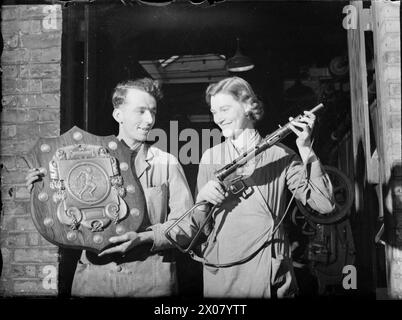 FROM MEDALS TO STEN GUNS : MUNITIONS PRODUCTION, BRITAIN, 1942 - deux ouvriers souriants d'une petite usine comparent un grand trophée sportif à un pistolet Sten. Cette usine avait auparavant produit des médailles, des plaques commémoratives et des trophées, mais a été remise à Sten production pendant la guerre Banque D'Images