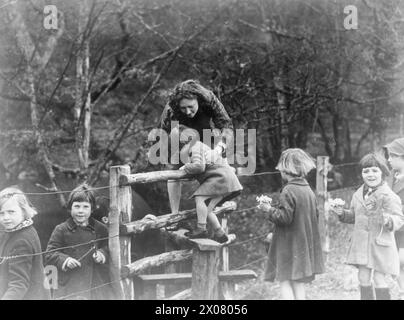 COLONIE POUR ARTISTES DE MOINS DE SIX ANS : ÉVACUÉS À DARTINGTON HALL, TOTNES, DEVON, ANGLETERRE, 1941. - L'enseignante Miss Betty Hall aide Marion Davison, 3 ans, la plus jeune évacuée à Dartington Hall, sur un stile dans le cadre de leur promenade dans la nature dans la campagne entourant le domaine de Dartington. D'autres évacués peuvent être vus sur la photo, beaucoup tiennent de petits bouquets de fleurs, qu'ils ont cueillis le long du chemin. Les arbres derrière eux offrent une toile de fond très différente des rues de Londres et Gravesend, Kent, qu'ils ont laissées derrière eux Banque D'Images