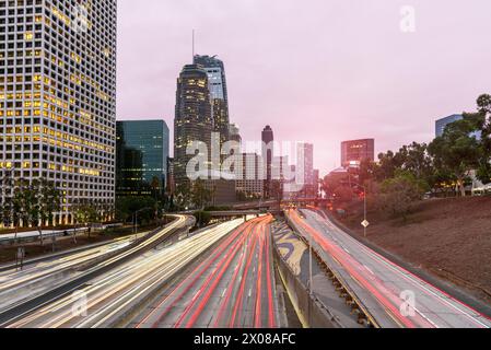 Longue exposition d'une autoroute très fréquentée bordant les gratte-ciel du centre-ville de Los Angeles au coucher du soleil en automne. Traînées de lumière. Banque D'Images