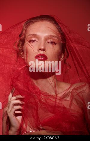 Une jeune femme respire la beauté classique tout en posant dans un studio dans une superbe robe rouge avec un voile en cascade sur sa tête. Banque D'Images