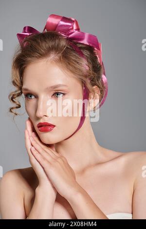 Une jeune femme avec un arc rose dans ses cheveux pose gracieusement dans un studio, exsudant la beauté classique sur un fond gris. Banque D'Images