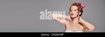 Une jeune femme exsudant la beauté classique tout en portant un arc rose sur sa tête, frappant une pose dans un cadre de studio sur un fond gris. Banque D'Images