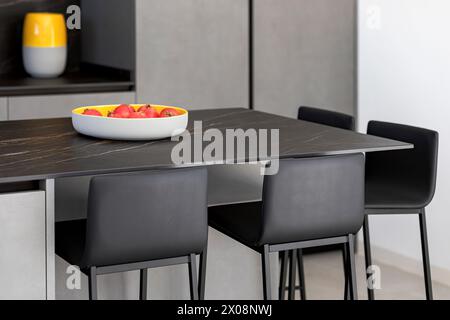 Une élégante salle à manger moderne avec une table sombre, des chaises noires et un bol de fruits lumineux comme pièce maîtresse Banque D'Images