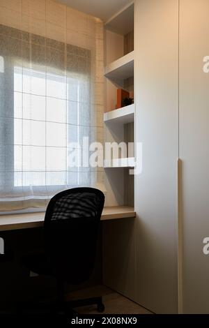 Installation moderne de bureau à domicile avec un bureau élégant, une chaise ergonomique et des étagères intégrées sous la lumière naturelle d'une fenêtre aux rideaux transparents Banque D'Images