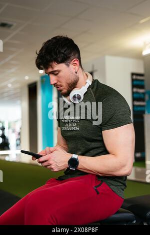 Jeune homme concentré avec des écouteurs à l'aide d'un smartphone dans la salle de gym. Banque D'Images