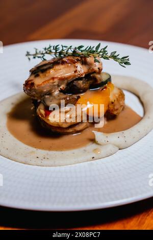 Médaillons de poulet superposés accompagnés de courgettes et de poivrons, recouverts d’une sauce crémeuse et de thym frais, présentés sur une assiette blanche texturée Banque D'Images