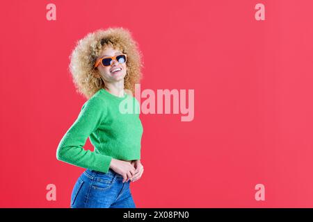 Une femme élégante avec un afro et des lunettes de soleil faisceaux avec confiance, portant un pull vert vif et un Jean bleu sur un fond rouge Banque D'Images