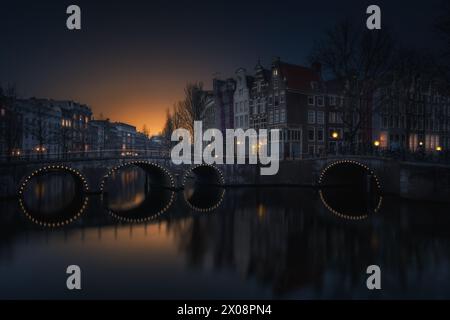 Les paisibles canaux d'Amsterdam se prélassent de la lueur du crépuscule, avec des bâtiments historiques et des ponts illuminés qui se reflètent dans les eaux calmes Banque D'Images