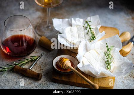 Fromage brie artisanal accompagné de miel, romarin frais et verres de vin rouge et blanc, le tout pour une dégustation raffinée. Banque D'Images
