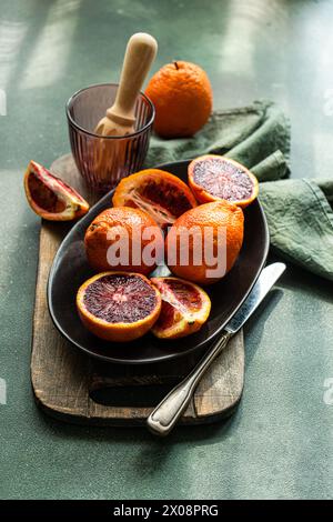 Un vif affichage d'oranges sanguines, certaines entières et d'autres coupées, sur une plaque sombre, avec un tissu vert et un alésoir d'agrumes en arrière-plan Banque D'Images