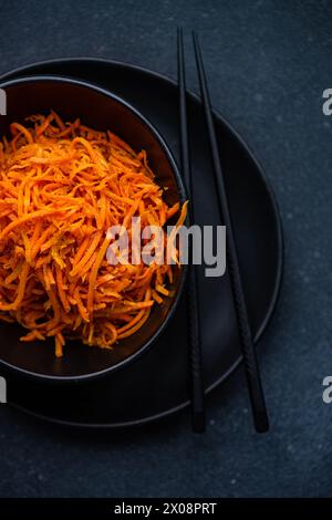 Une image verticale capturant un gros plan d'une salade de carottes fraîches râpées servie dans un élégant bol noir avec une paire de baguettes noires sur un fond sombre Banque D'Images