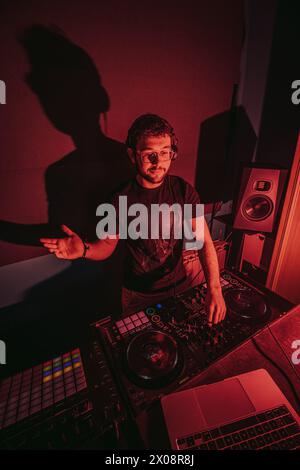 Un DJ masculin concentré avec des écouteurs mixe la musique sur sa terrasse sous un éclairage ambiant rouge, exsudant des vibrations fraîches lors d'une soirée Banque D'Images