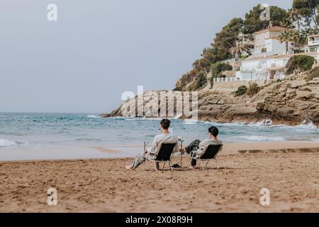 Vue arrière de deux personnes se prélassant dans des chaises sur une plage de sable avec des vagues et la côte rocheuse en arrière-plan. Banque D'Images