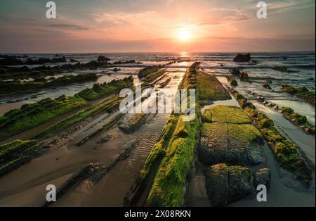 Le soleil couchant baigne Playa de Barrika dans une lumière dorée, soulignant les roches moussues texturées qui mènent à la mer embrassée par le soleil Banque D'Images