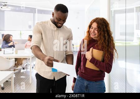 Homme noir et femme blanche discutant sur un document dans un cadre de bureau moderne, souriant et regardant le document. Banque D'Images