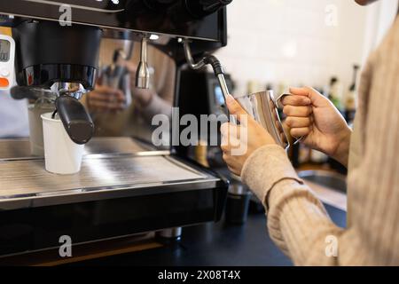 Gros plan des mains d'un barista qui fait cuire du lait à la vapeur dans un pichet métallique avec une machine à espresso professionnelle dans un café. Banque D'Images