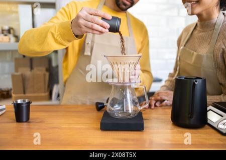 Un barista vêtu d'un pull jaune verse du café moulu dans une machine à verser pendant que son collègue regarde avec intérêt, dans un cadre de café confortable. Banque D'Images