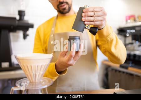 Un barista dans un tablier jaune moud des grains de café frais à l'aide d'un moulin manuel dans une tasse portable dans un café moderne. Banque D'Images