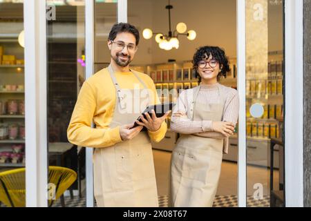 Deux entrepreneurs souriants dans des tabliers se tiennent à l’entrée de leur café, prêts à accueillir les clients. Banque D'Images