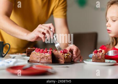 Une main adulte coupée méconnaissable coupe un gâteau au chocolat orné de baies alors qu'un enfant dans un dessus rouge regarde, anticipant une tranche. Banque D'Images