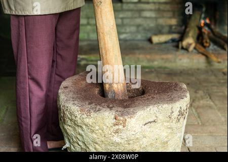Une personne rognée méconnaissable dans des vêtements traditionnels moud manuellement les grains de café dans un mortier de pierre avec un grand pilon en bois, avec un fond o Banque D'Images