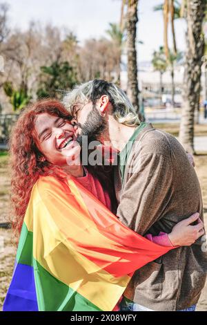 Un couple enveloppé dans un drapeau arc-en-ciel partage une tendresse étreinte, leur rire et leur affection brillent à travers, avec des palmiers en arrière-plan Banque D'Images