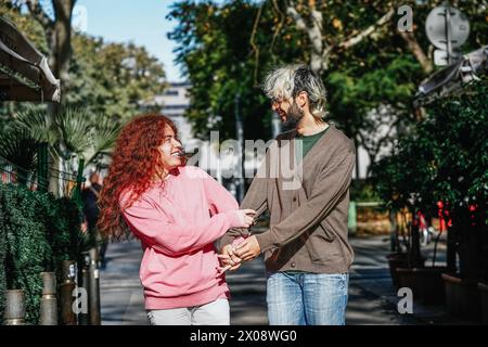 Un couple partage un moment ludique et affectueux, se tenant la main et riant sur une passerelle ensoleillée de la ville, bordée de verdure luxuriante Banque D'Images