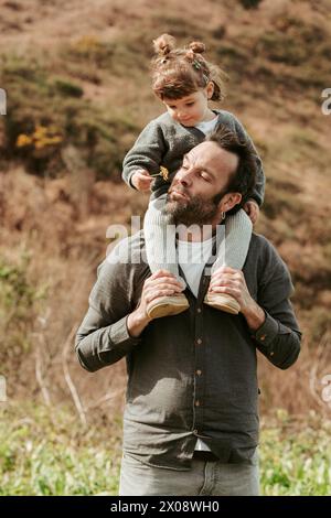 Un moment d'amour comme un père porte sa jeune fille sur ses épaules à l'extérieur, avec un doux sourire sur son visage alors qu'elle examine une fleur Banque D'Images