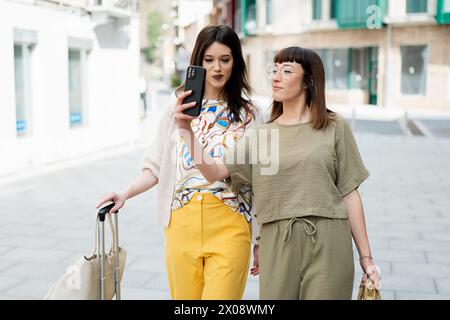 Deux femmes soucieuses de la mode marchant sur un trottoir de la ville, l'une tenant un smartphone et un sac à provisions, toutes deux arborant une tenue printanière tendance Banque D'Images
