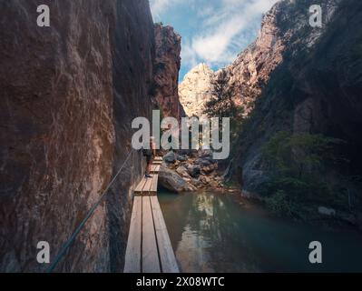 Un randonneur explore un sentier de montagne pittoresque le long d'une promenade en bois à côté d'une rivière tranquille, entourée de falaises imposantes Banque D'Images