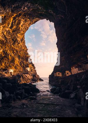 Une arche naturelle à couper le souffle s'élevant au-dessus d'une crique rocheuse avec des maisons blanches nichées contre la falaise sur l'île de la Palma, en Espagne, tandis que le coucher du soleil illumine Banque D'Images