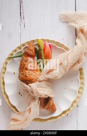 Un croissant sur le thème de Pâques enveloppé d'un tissu rustique, orné d'une tulipe vibrante, présenté sur une assiette décorative, invoquant l'essence du printemps Banque D'Images