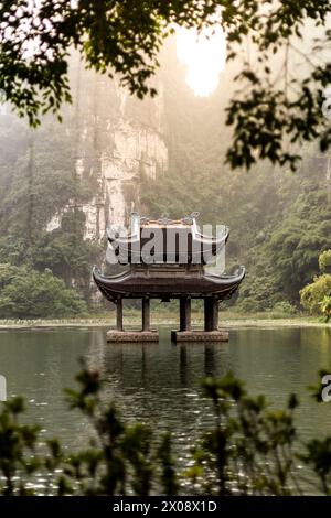 Une pagode tranquille se dresse sur pilotis au-dessus d'un lac calme, encadrée par des falaises de calcaire karstiques et un feuillage luxuriant au Vietnam, évoquant un sentiment de paix et d'esprit Banque D'Images