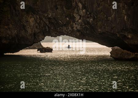 Un bateau traditionnel avec pêcheur méconnaissable glisse à travers les eaux sereines de la baie d'Halong, Vietnam, en passant sous une arche de calcaire naturelle avec Banque D'Images