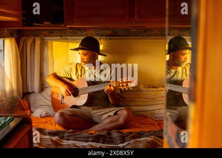 Un musicien masculin joue de la guitare classique à l'intérieur de son camping-car confortable, immergé dans la créativité tandis que la lueur chaude du coucher de soleil se reflète sur la mer. Banque D'Images