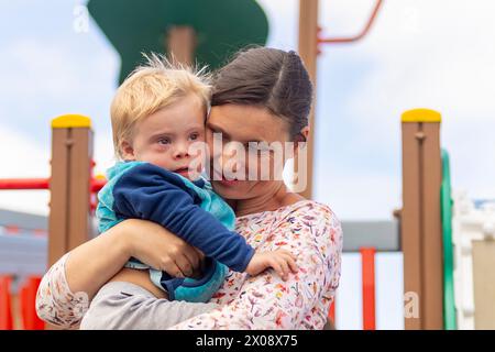 Tendresse étreinte d'une mère tenant son enfant atteint du syndrome de Down sur une aire de jeux, capturant un moment de pur amour et de soins. Banque D'Images