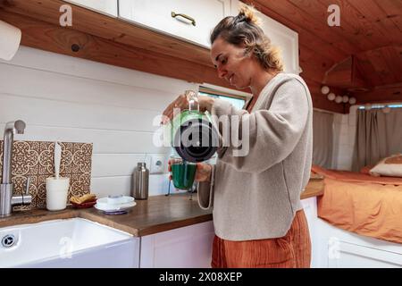 Femme verse une boisson à l'intérieur d'une fourgonnette bien équipée, représentant l'espace de vie d'une famille toute l'année avec une cuisine intégrée et le confort de la maison. Banque D'Images