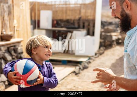 Un garçon souriant tenant un ballon regarde son père dans un espace de vie extérieur ouvert, reflétant la vie de famille dans un van avec une terrasse et un jardin faits maison. Banque D'Images